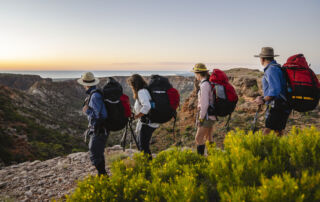 Four walkers with their large hiking backpacks looking out at the pink and orange hues of Western Australia's horizon. Image supplied by Destination WA.